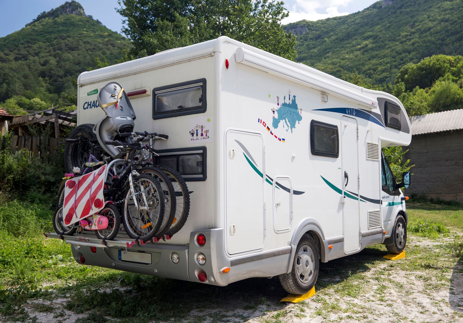 An RV with bicycles attached in a scenic outdoor setting, surrounded by mountains and greenery.